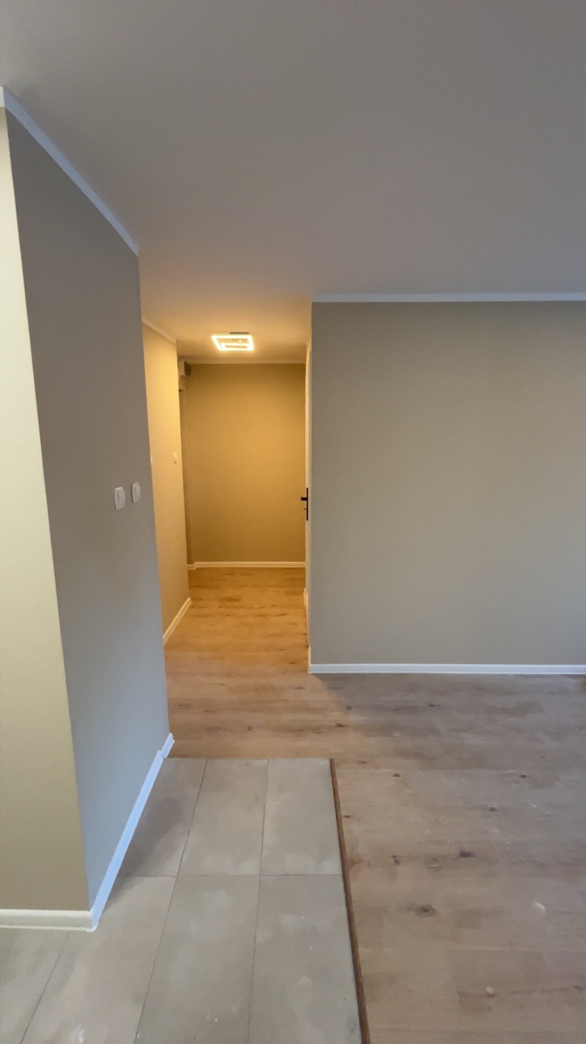 Beżowe ściany po malowaniu w pustym pokoju z fragmentem ułożonej podłogi z płytek i drewnianych paneli. Widok na korytarz z oświetleniem sufitowym.