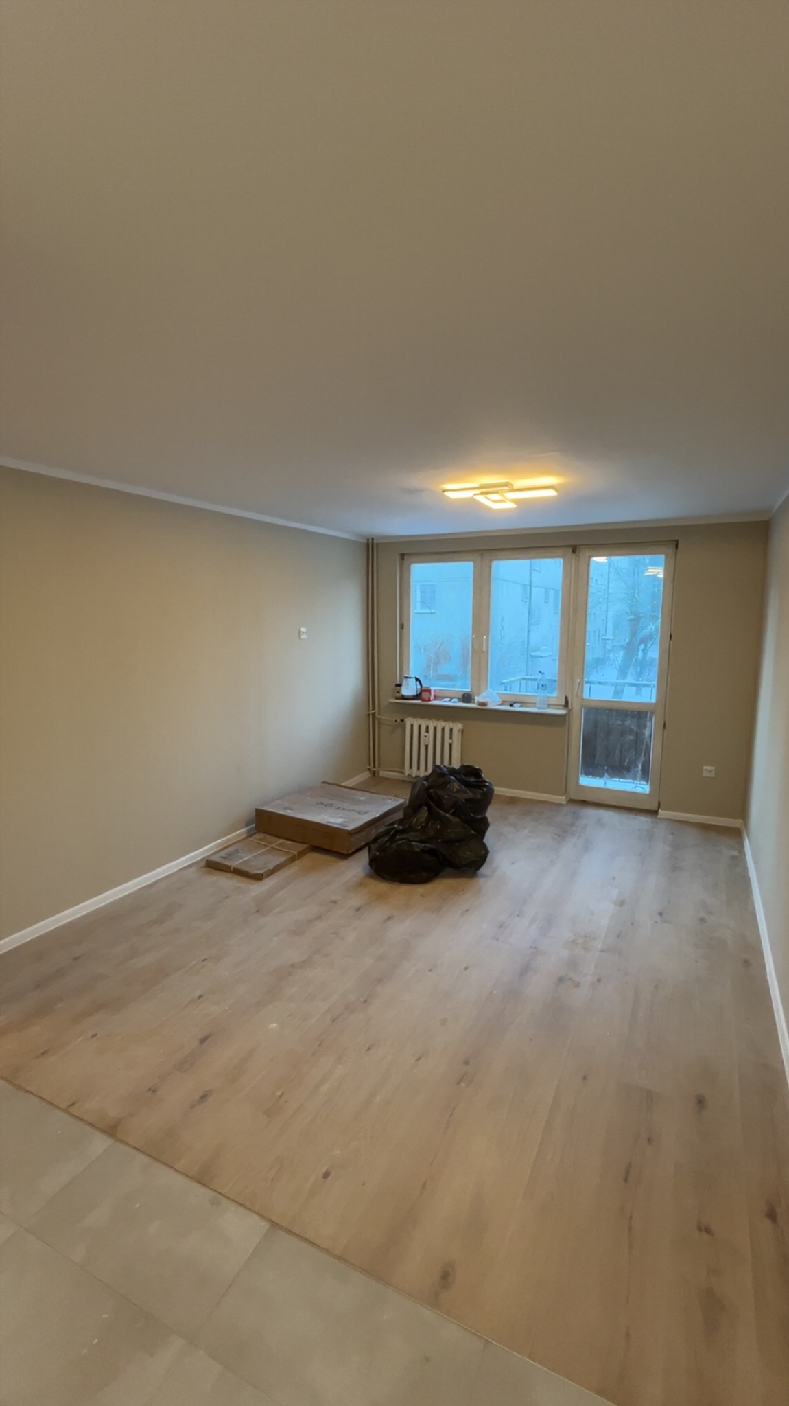 Pokój po remoncie podłogi z panelami, widoczne worki z odpadami i kartony po panelach. Ściany pomalowane na beżowo, okno balkonowe z widokiem na zewnątrz.