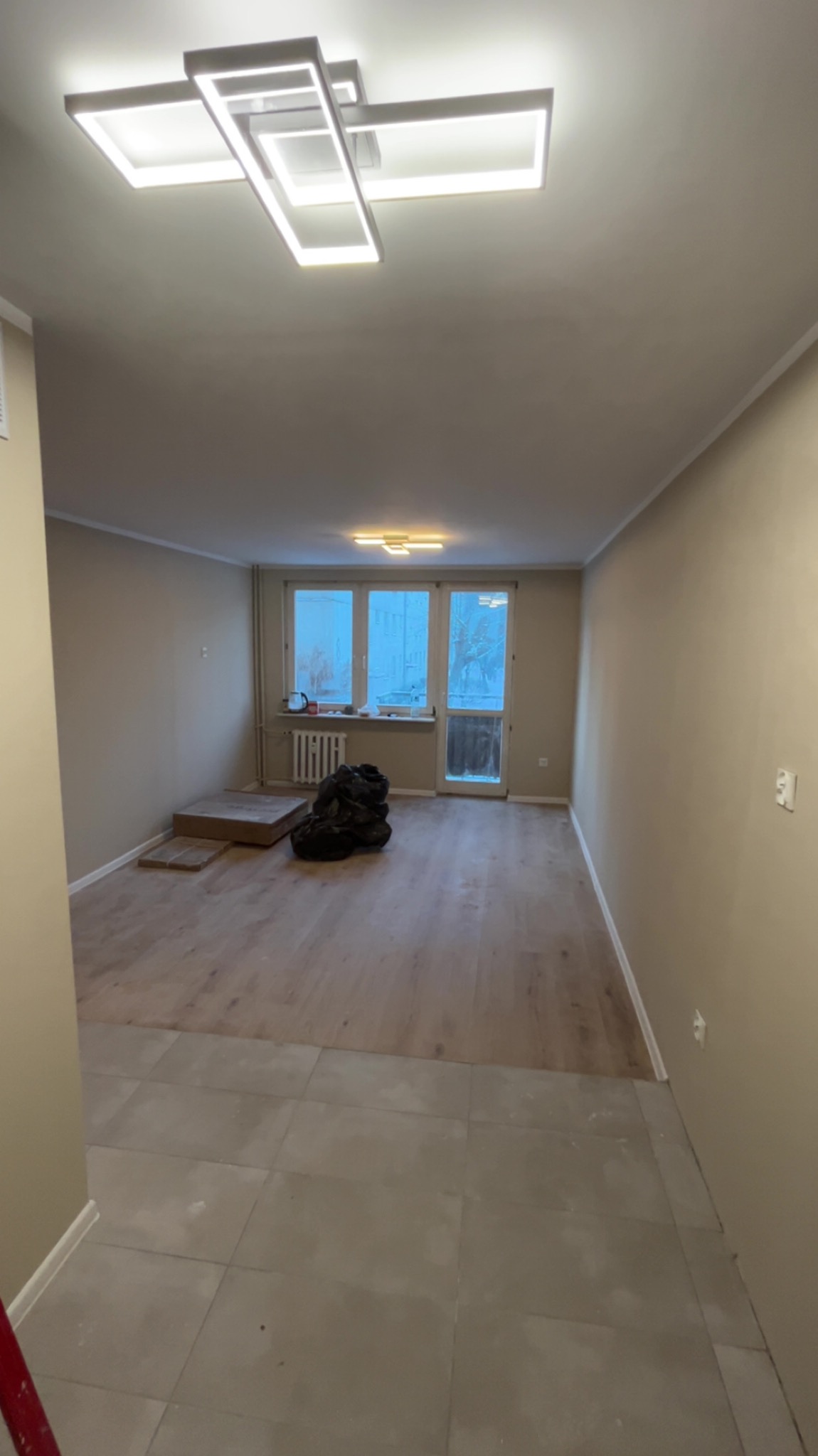 Jasne wnętrze po remoncie: świeżo pomalowane ściany w odcieniach beżu, nowa podłoga z paneli, nowoczesna lampa sufitowa. Widoczne ślady prac wykończeniowych.