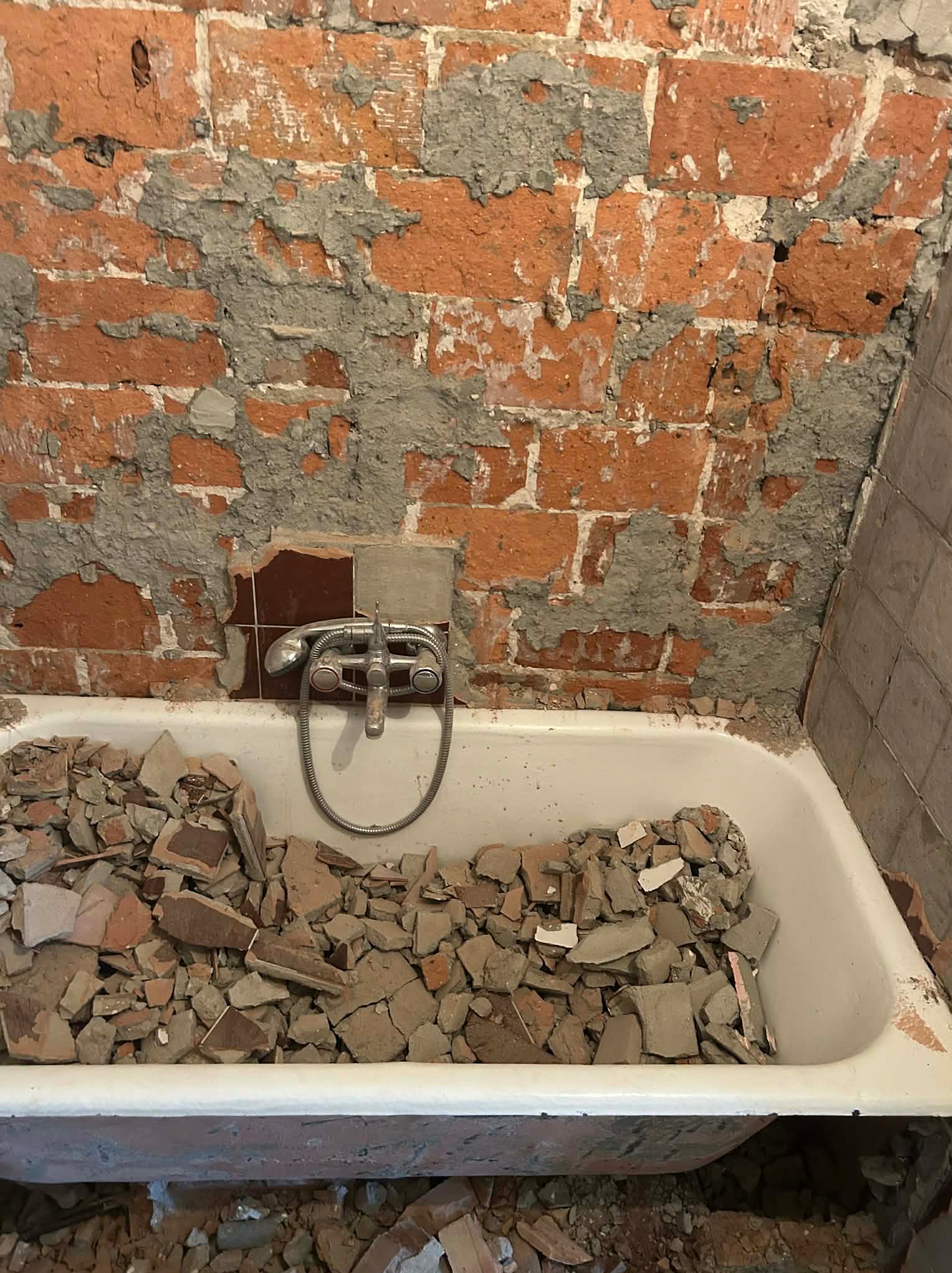Demontaż płytek w łazience: gruz w wannie, odsłonięta cegła na ścianie, widoczna armatura. Etap przygotowania do remontu, widok z góry.