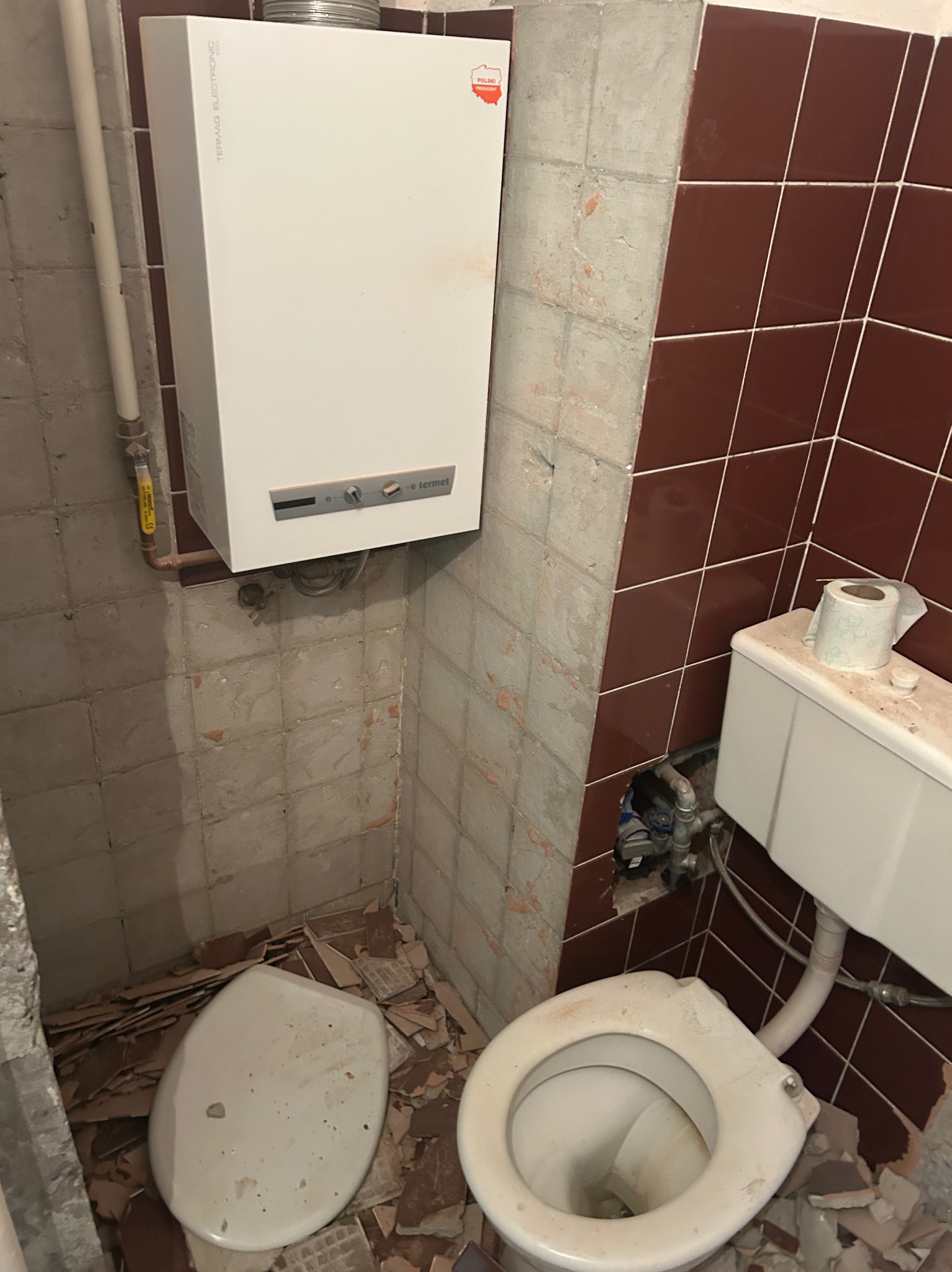 Demontaż starej łazienki: wiszący bojler Termet, częściowo zdemontowane brązowe płytki, muszla klozetowa i deska na podłodze pokrytej gruzem.