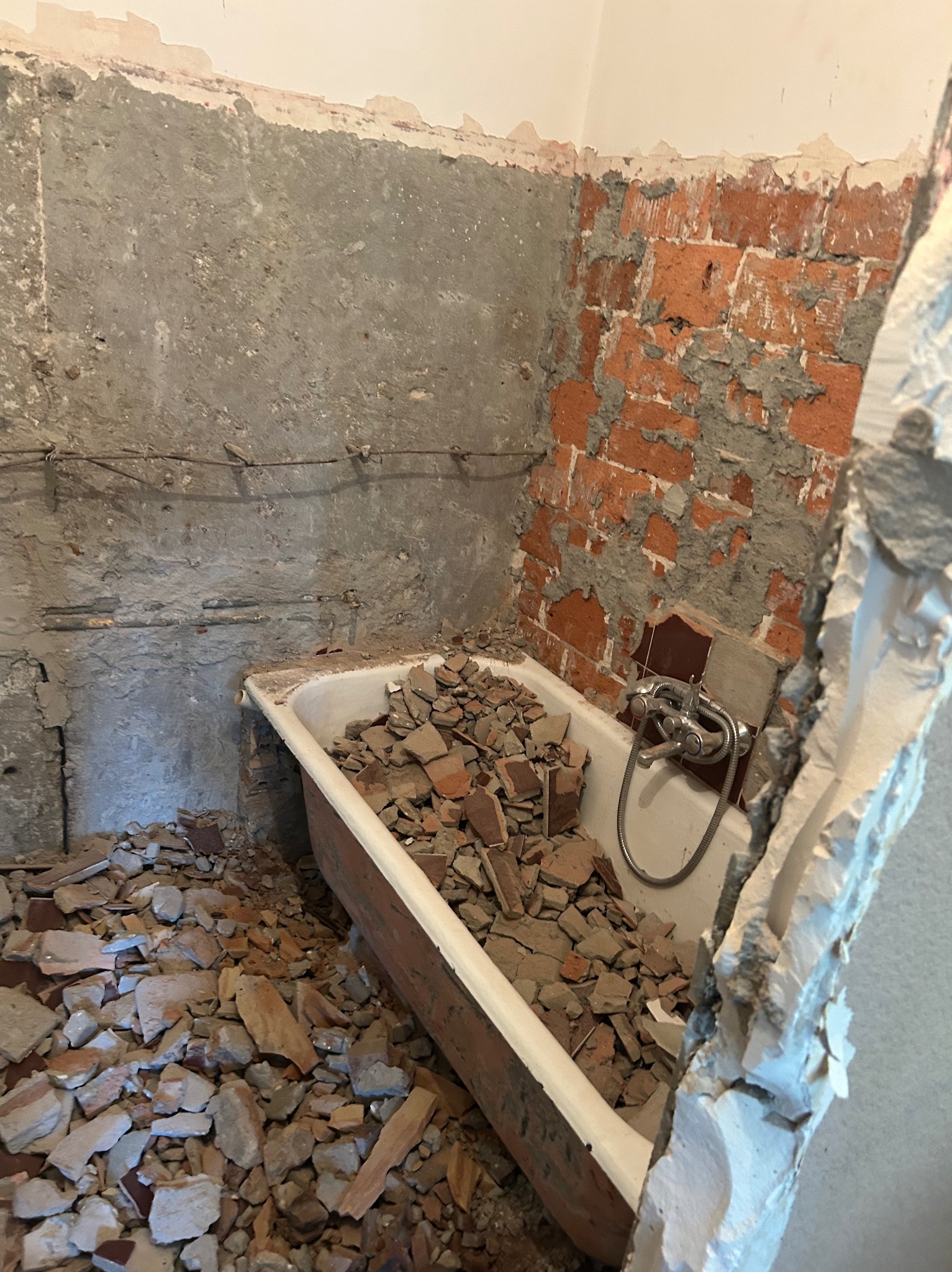 Demontaż płytek w łazience: gruz w wannie, odsłonięta cegła i surowy beton na ścianach. Widoczne rury i armatura. Etap przygotowania do remontu.