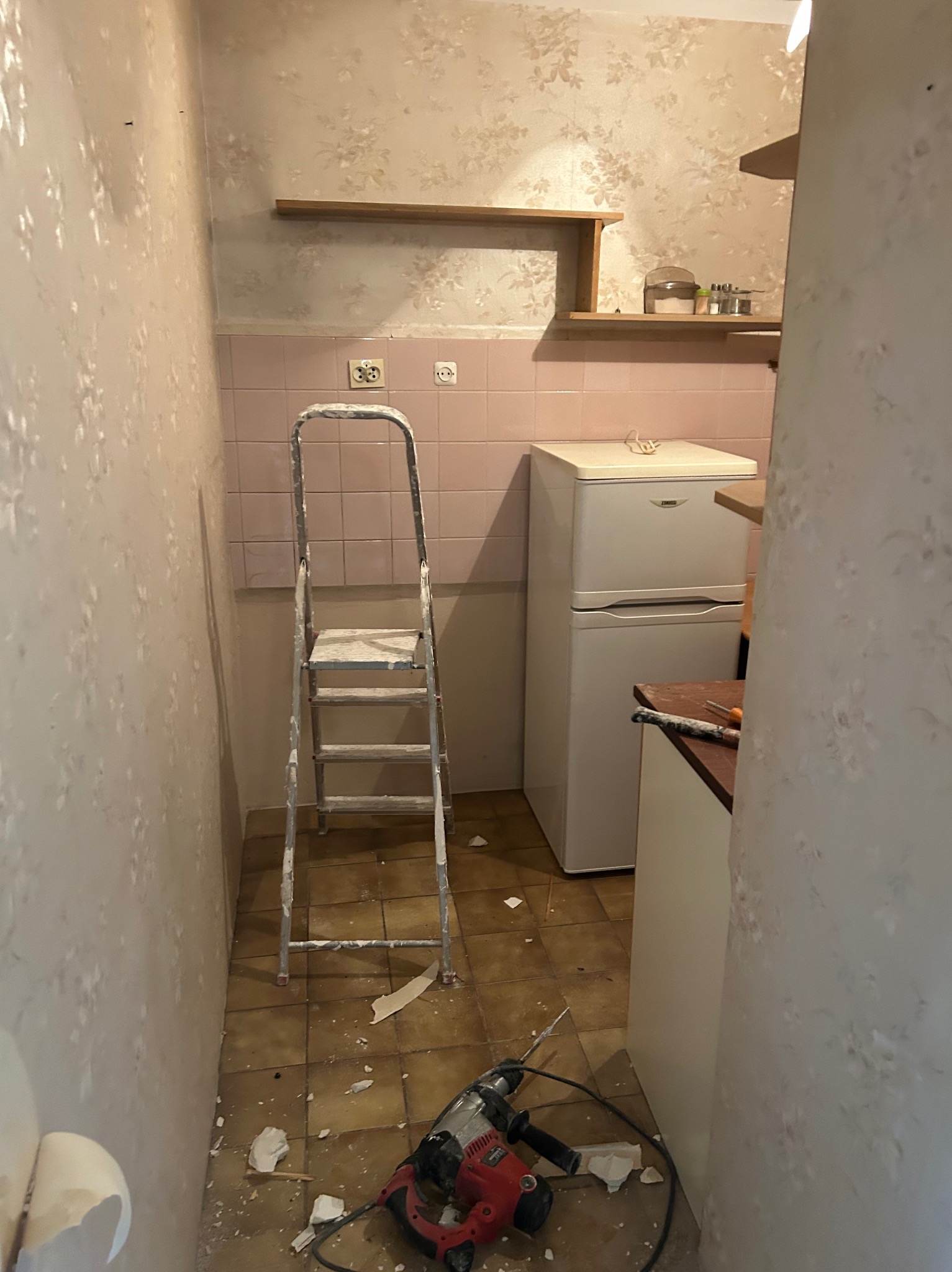 Wnętrze kuchni w trakcie remontu: drabina malarska, ślady po skuciu tynku na ścianach, różowa glazura, sprzęt AGD i narzędzia (wiertarka udarowa) na podłodze.