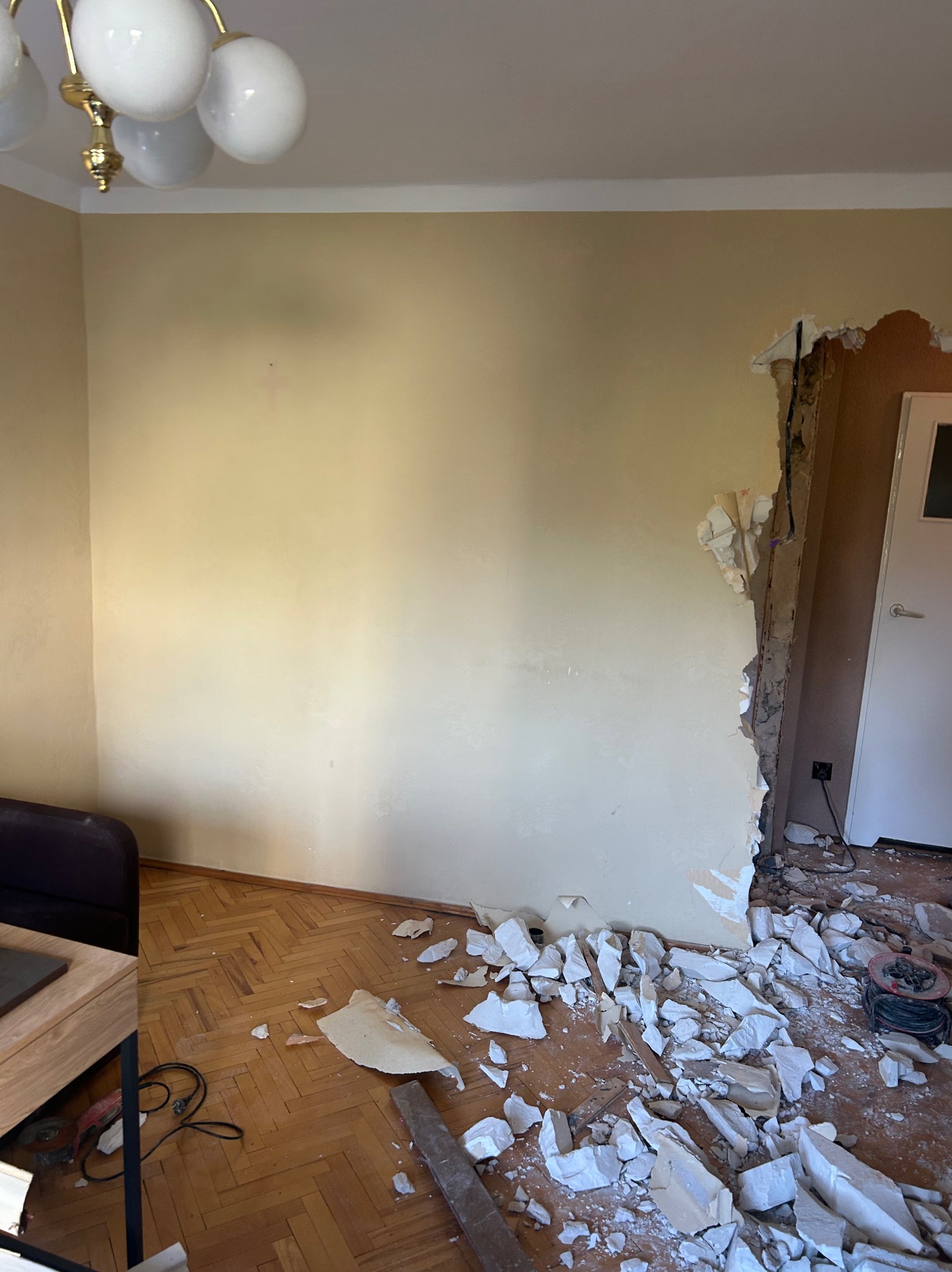Demontaż ściany działowej: fragment ściany usunięty, widoczne instalacje, gruz na parkiecie. Przygotowanie do remontu, widoczna lampa i fragment mebli.