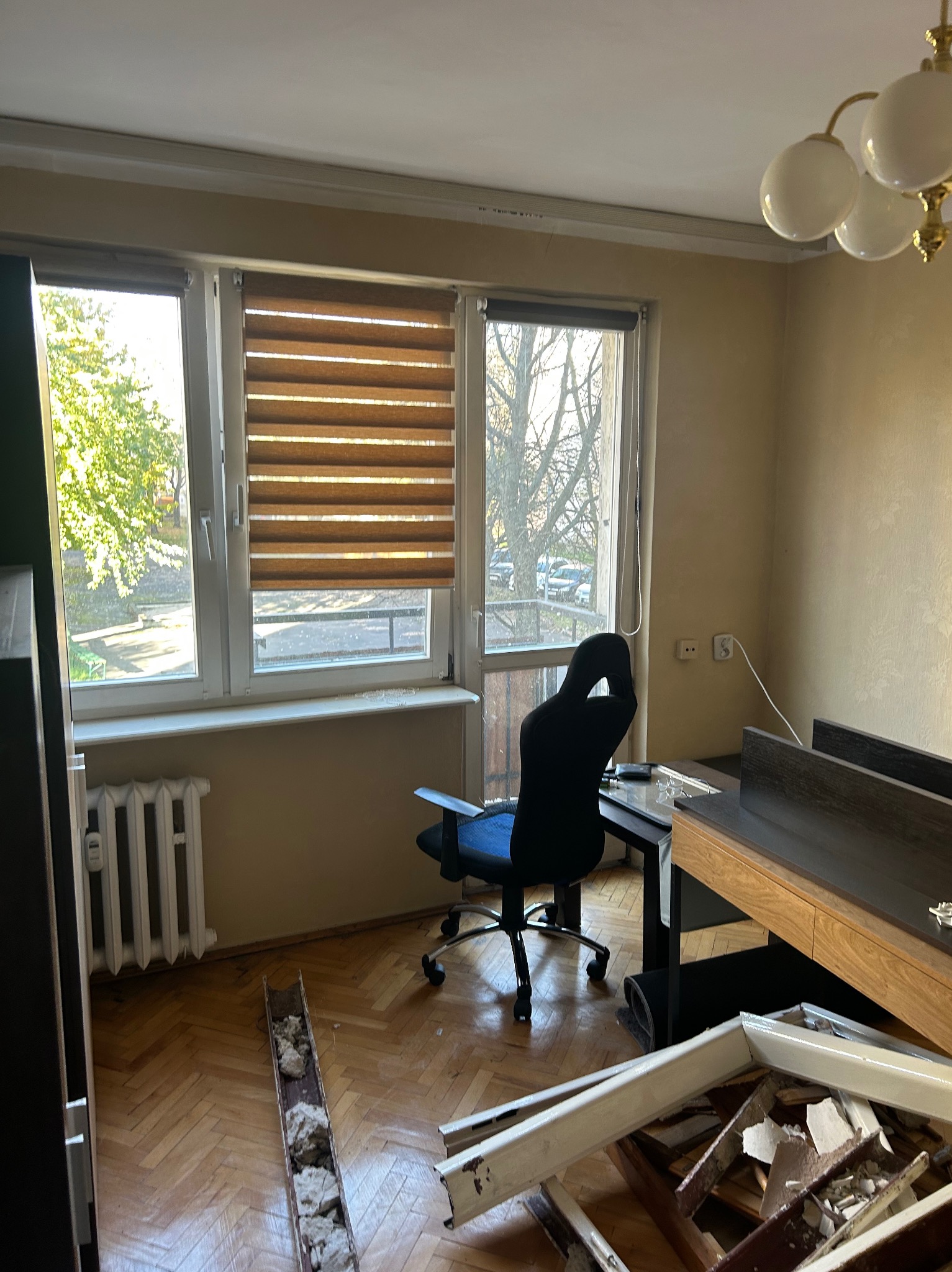 Pokój w trakcie demontażu: widoczne okna z roletami, biurko, krzesło i gruz po demontażu. Parkiet na podłodze. Widok na zieleń za oknem.