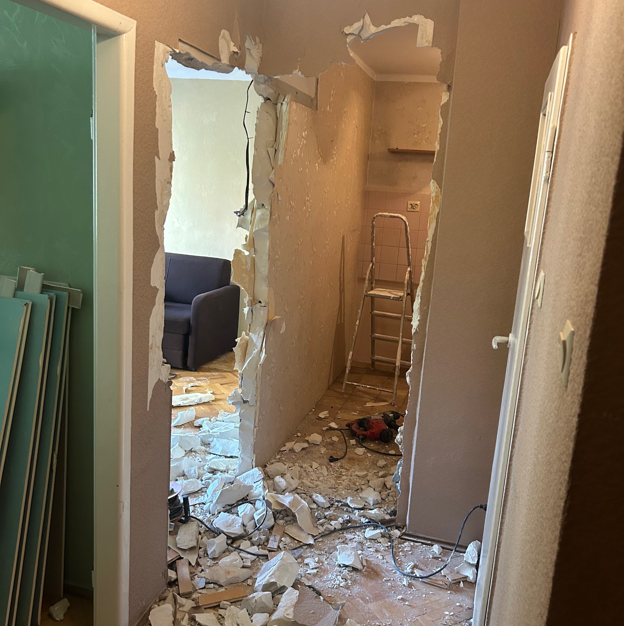 Demontaż ściany działowej z widocznym gruzem, narzędziami i fragmentami instalacji elektrycznej. W tle widoczny pokój z sofą i drabina w pomieszczeniu z różowymi płytkami.