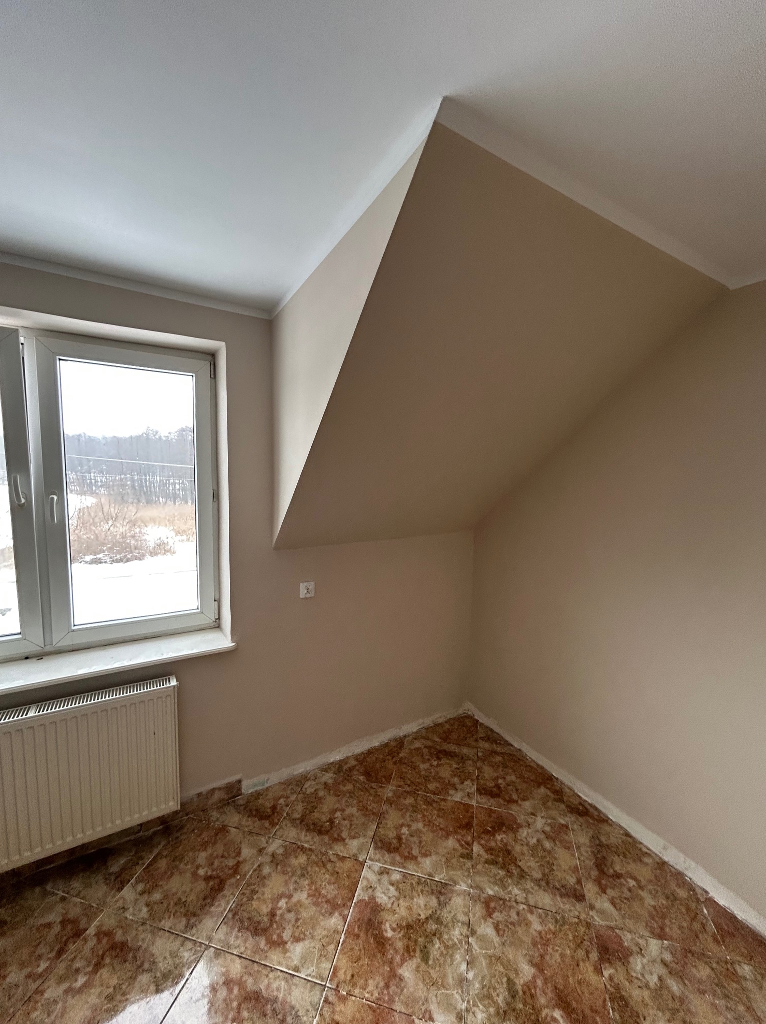 Pokój na poddaszu z oknem, brązowymi płytkami podłogowymi i beżowymi ścianami. Widoczny grzejnik pod oknem i skośny sufit. Neutralne kolory, prosta aranżacja.