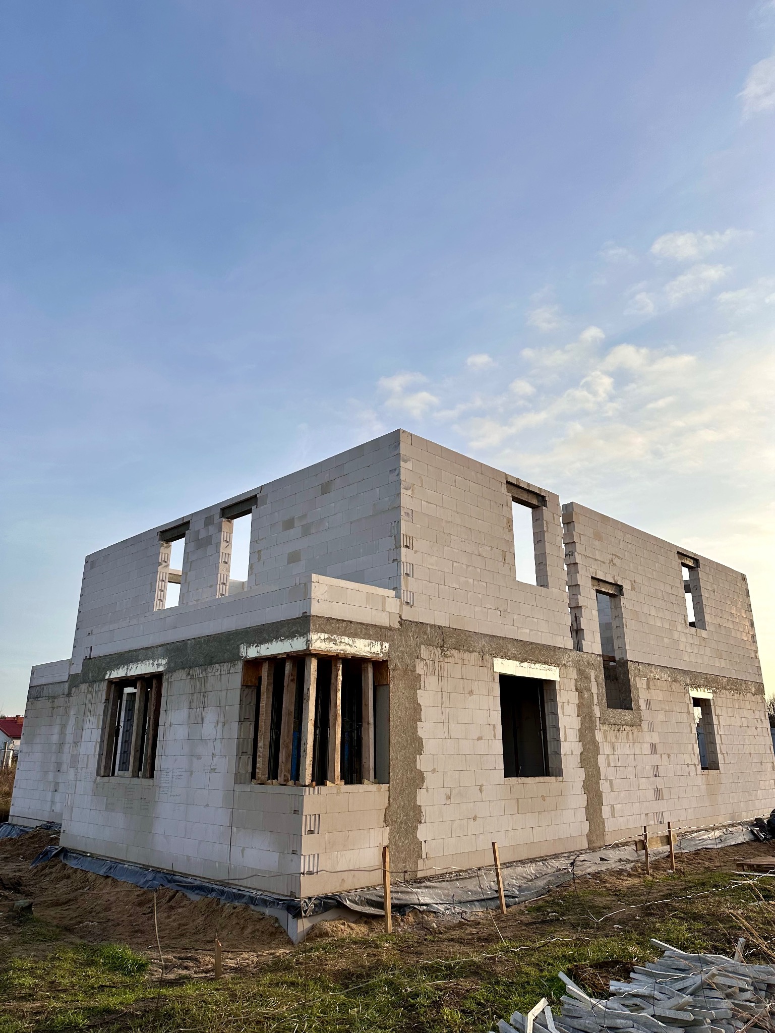 Wznoszenie ścian domu z betonu komórkowego. Widoczne otwory okienne i drzwiowe, drewniane podpory. Budowa w toku, surowy stan. Błękitne niebo w tle.