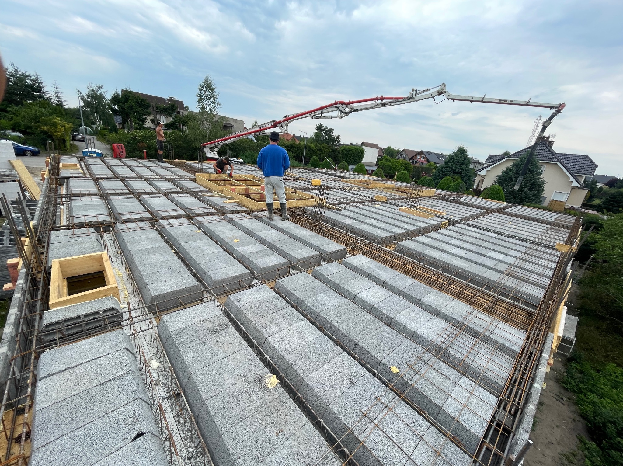 Budowa stropu Teriva z użyciem pompy do betonu. Widoczne bloczki, zbrojenie i pracownicy na budowie. Ujęcie z góry, w tle zabudowa jednorodzinna.
