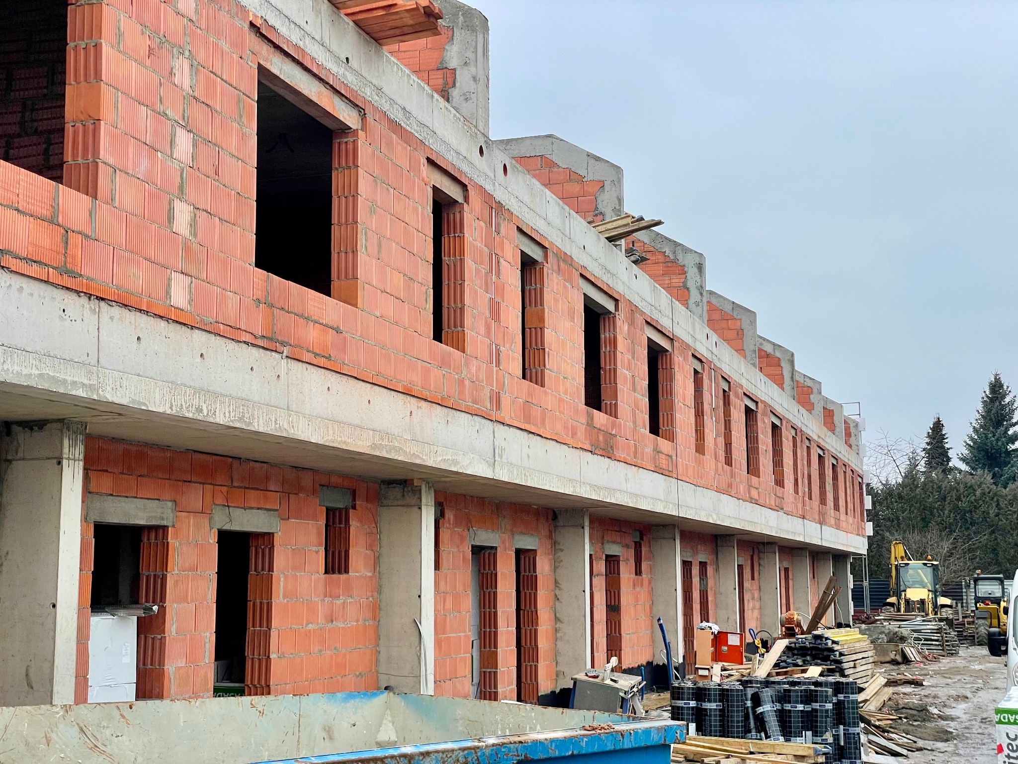 Wznoszenie ścian z czerwonej cegły ceramicznej na budowie szeregowca. Widoczne otwory okienne i betonowe nadproża. W tle sprzęt budowlany i drzewa.