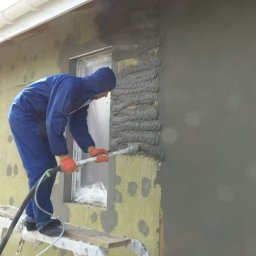 ECO PRO Ivan Pylyp - Pracownik w kombinezonie nanosi tynk maszynowy na ocieploną ścianę budynku z użyciem lancy, stojąc na aluminiowym podeście. Część ściany jest już otynkowana.