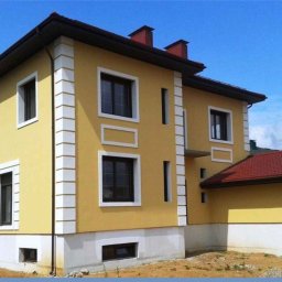 ECO PRO Ivan Pylyp - Dwukondygnacyjny dom z żółtą elewacją i białymi zdobieniami okiennymi, widoczny z boku, na tle błękitnego nieba. Czerwony dach i częściowo wykończony teren.