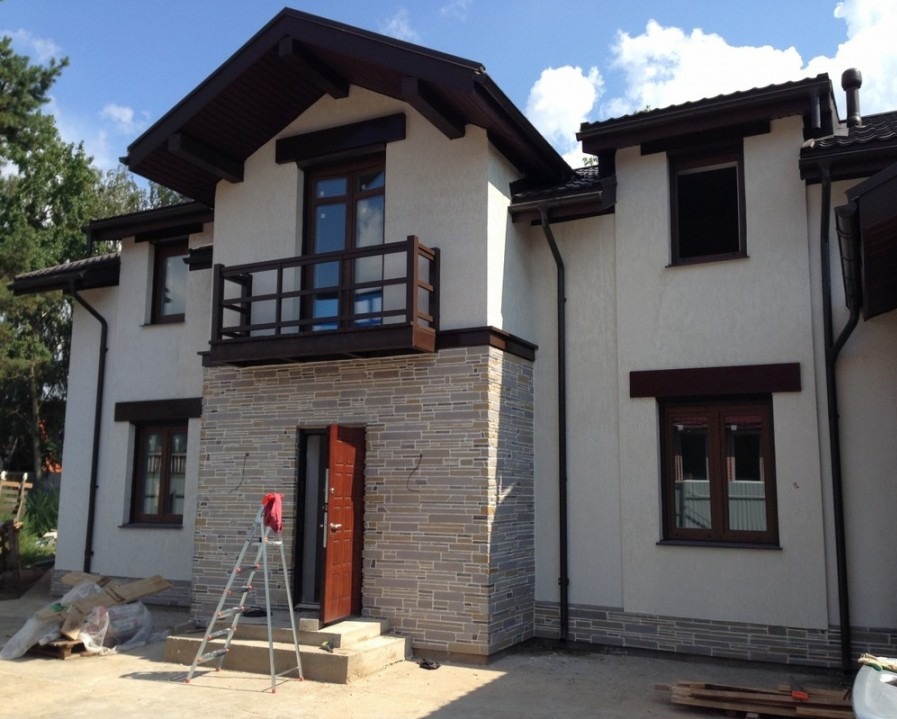 Dwukondygnacyjny dom z balkonem, wykończony jasnym tynkiem i kamieniem elewacyjnym. Otwarta, brązowa stolarka okienna i drzwiowa. Drabina oparta o wejście.