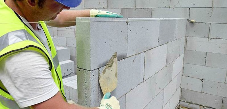 Murarz w odblaskowej kamizelce, z kielnią w dłoni, muruje ścianę z białych bloczków. Widoczna linka murarska i zapas bloczków.