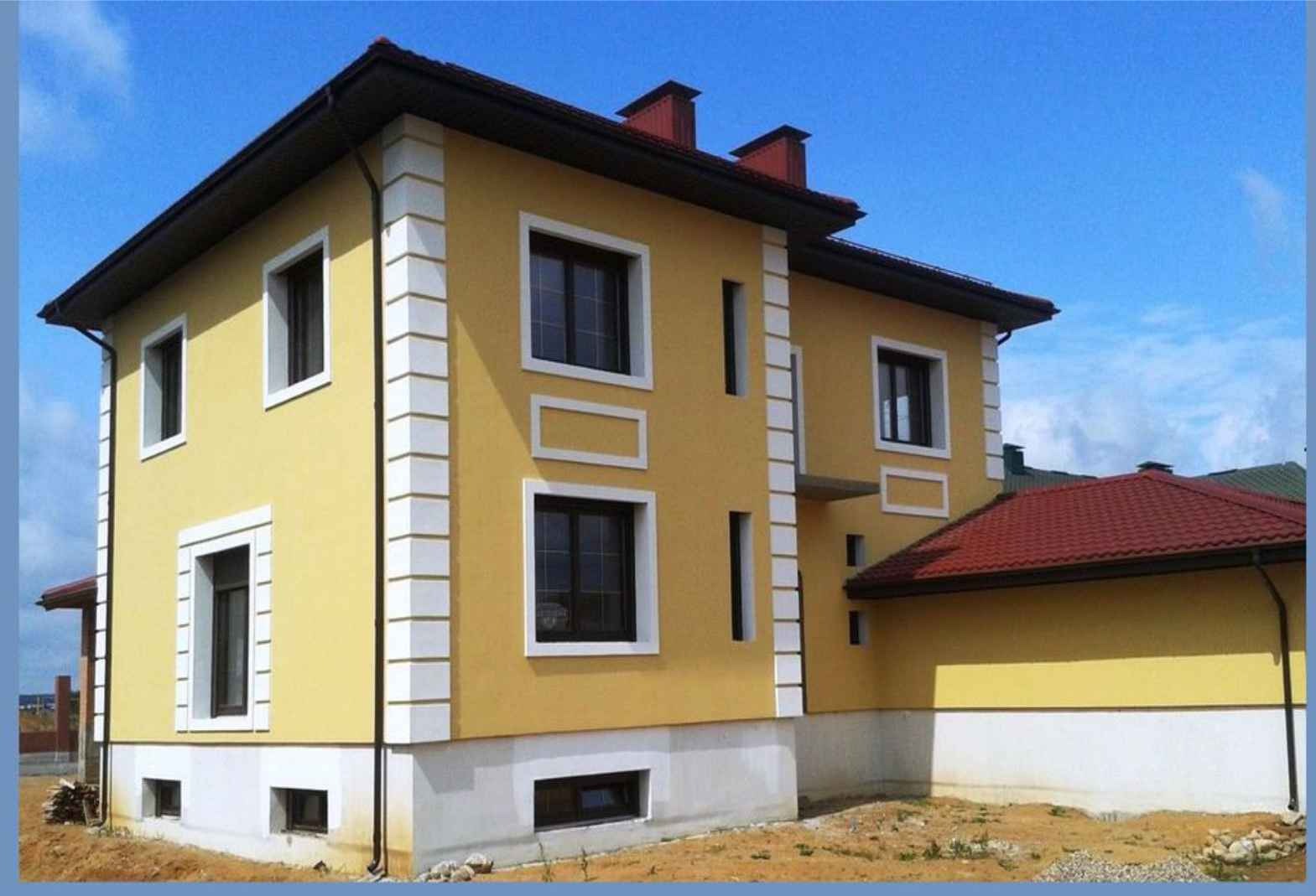 Dwukondygnacyjny dom z żółtą elewacją i białymi zdobieniami okiennymi, widoczny z boku, na tle błękitnego nieba. Czerwony dach i częściowo wykończony teren.