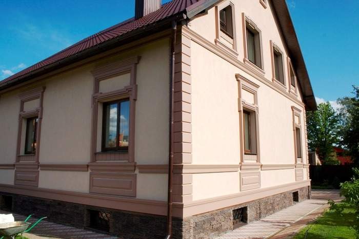 Elewacja domu jednorodzinnego z dekoracyjnymi elementami architektonicznymi wokół okien i na cokole, w ciepłych odcieniach beżu i brązu, z widocznym fragmentem ogrodu.