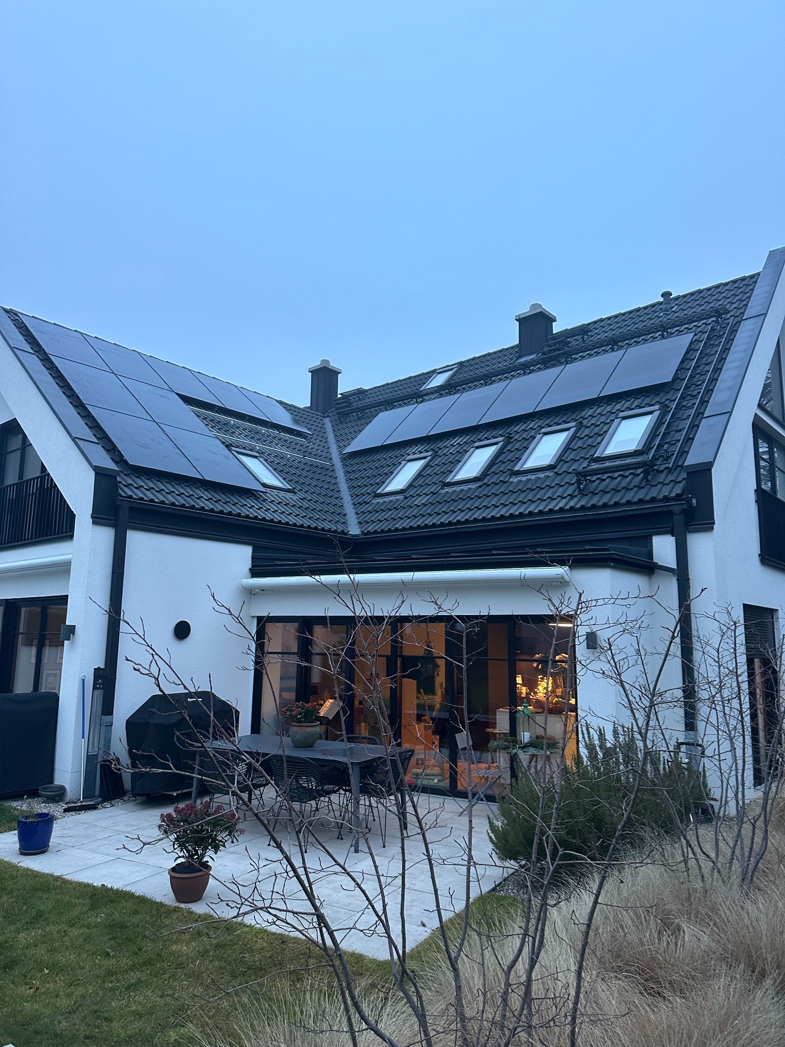 Biały dom z czarnym dachem i panelami słonecznymi. Widoczne okna dachowe, taras z meblami ogrodowymi i roślinami. Ujęcie z dołu, pochmurne niebo.