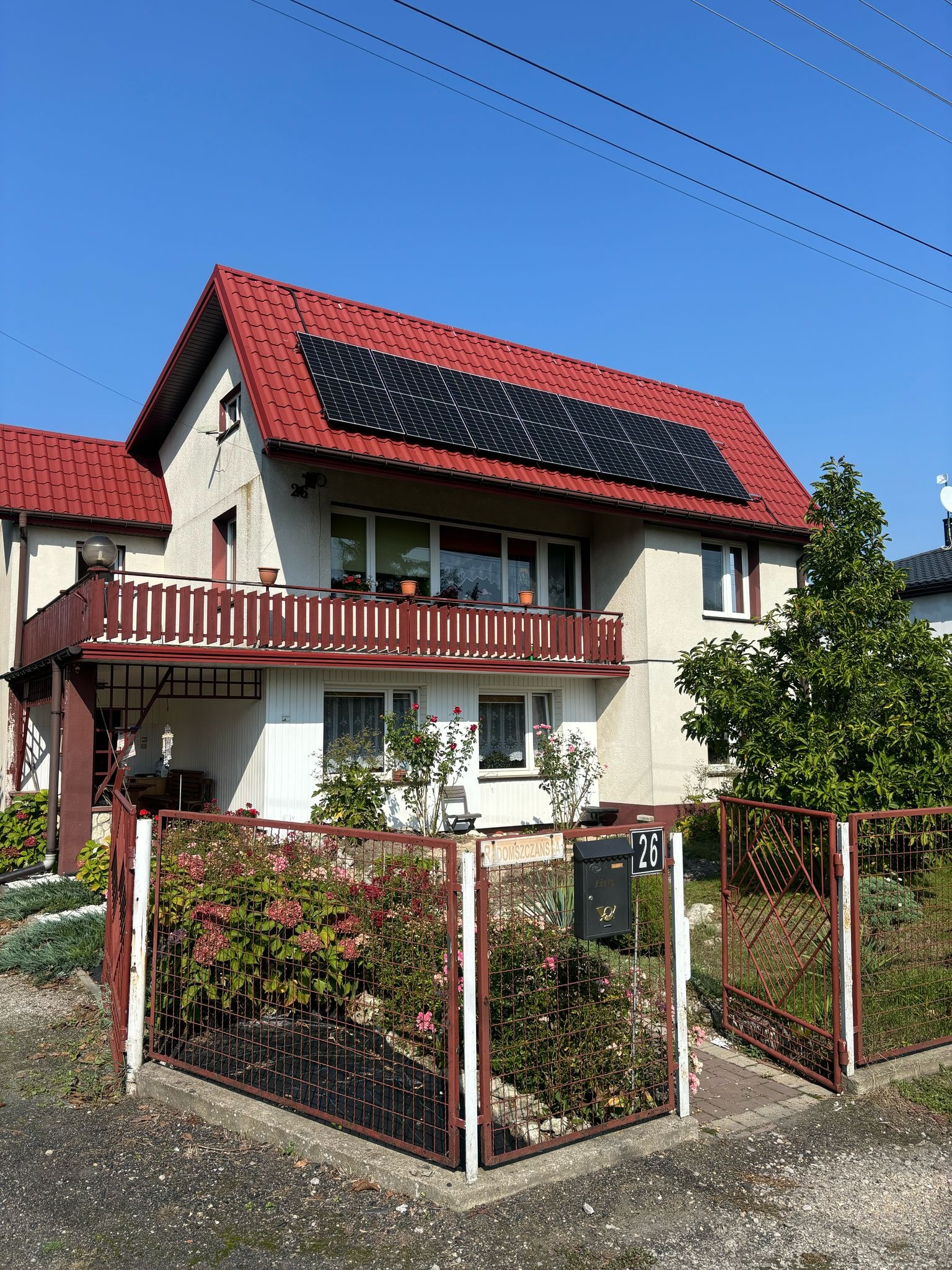 Dom jednorodzinny z czerwonym dachem i panelami fotowoltaicznymi na dachu. Widoczny balkon z drewnianą balustradą i ogród z metalowym ogrodzeniem.