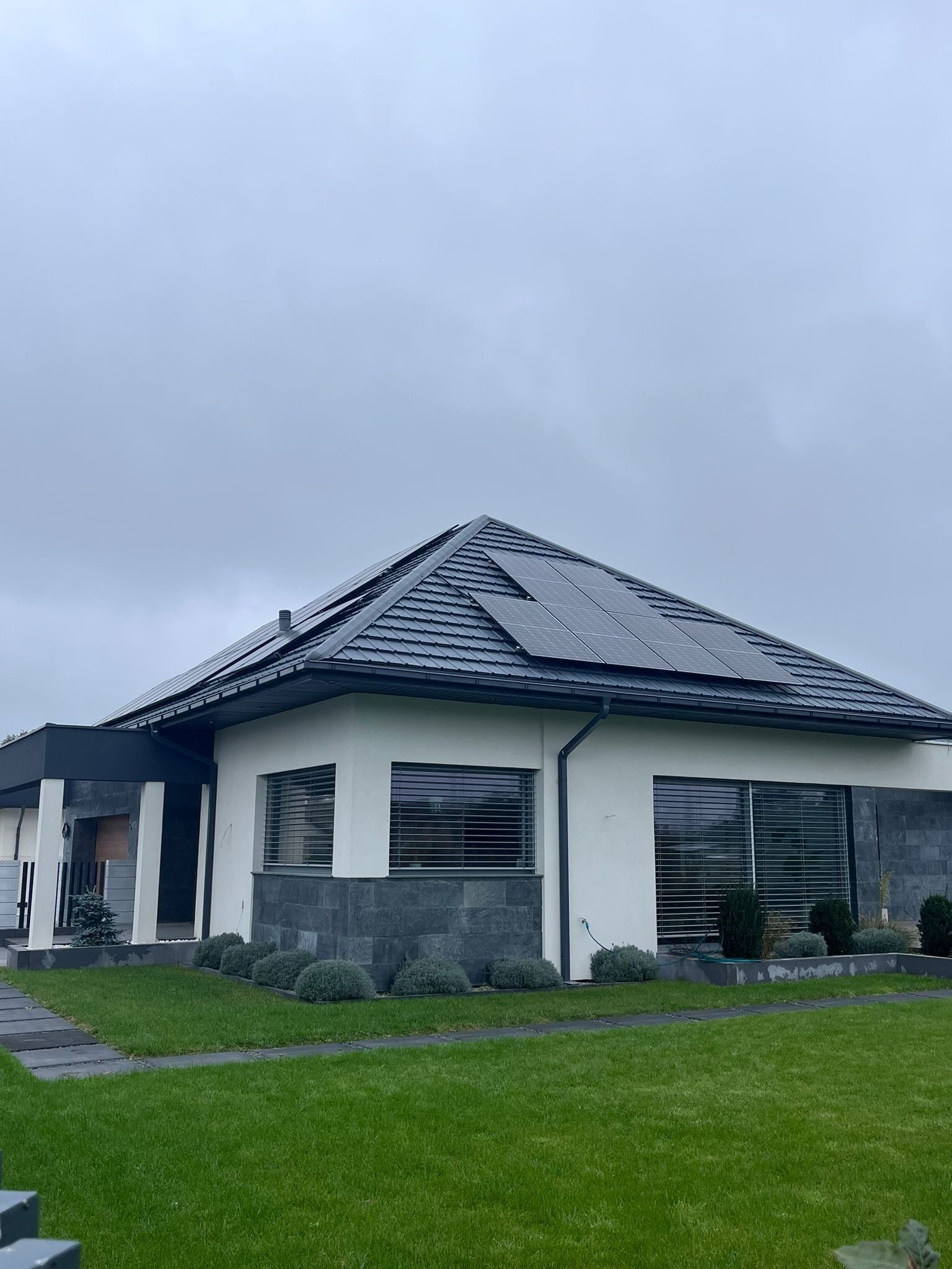 Nowoczesny dom jednorodzinny z panelami fotowoltaicznymi na dachu, otoczony zielonym trawnikiem i ozdobnymi krzewami w pochmurny dzień.