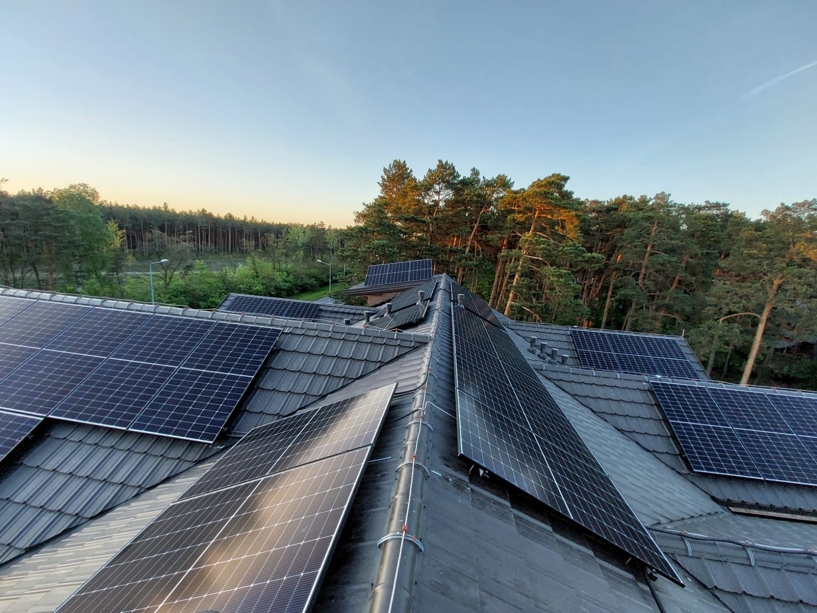 Panele fotowoltaiczne na dachu z szarej dachówki, widok z góry, w tle las iglasty oświetlony zachodzącym słońcem. Instalacja paneli na skośnym dachu.