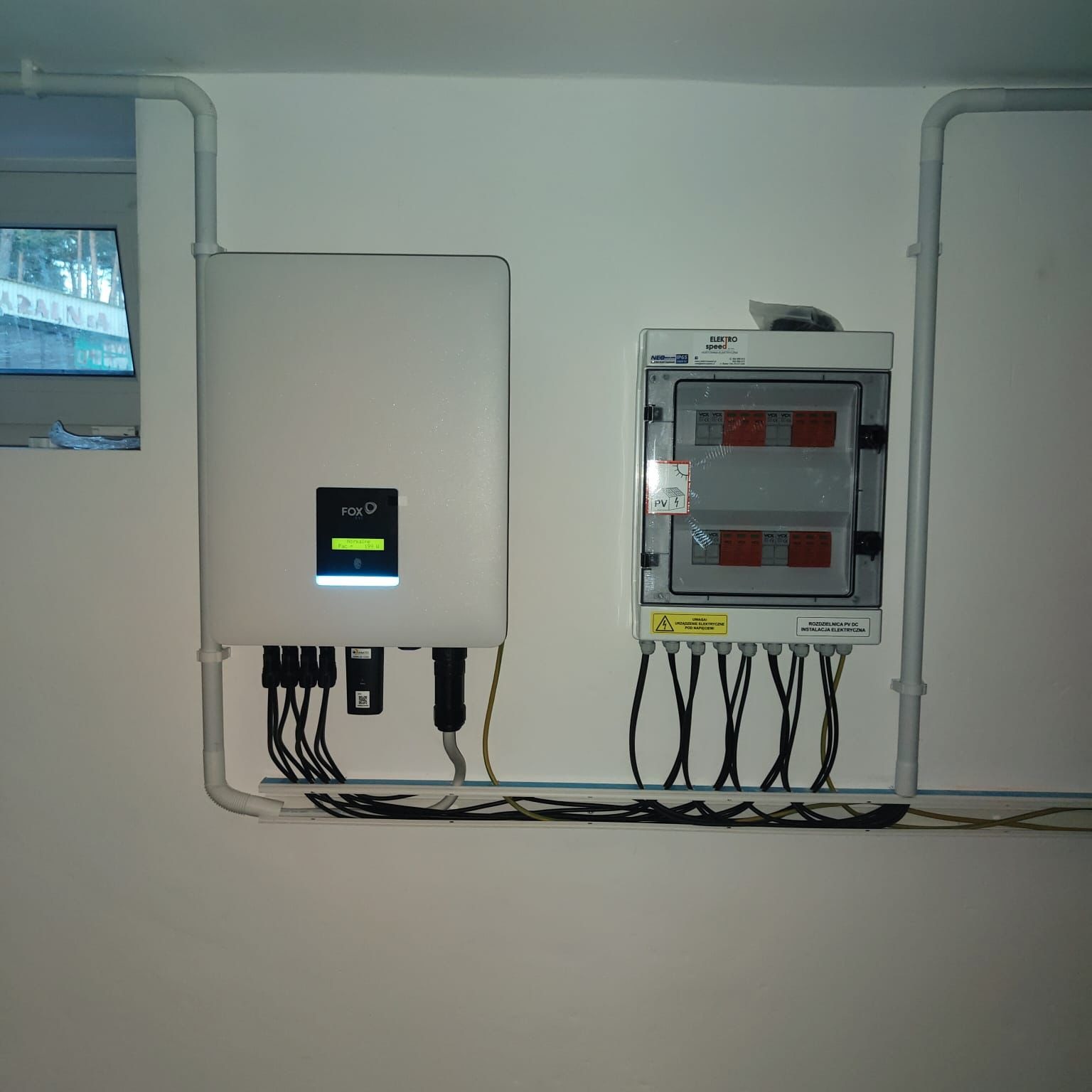 Biały inwerter Fox połączony z rozdzielnicą PV DC Elektro Speed na białej ścianie, z widocznymi przewodami i rurami instalacyjnymi. Widok z bliska, wnętrze.