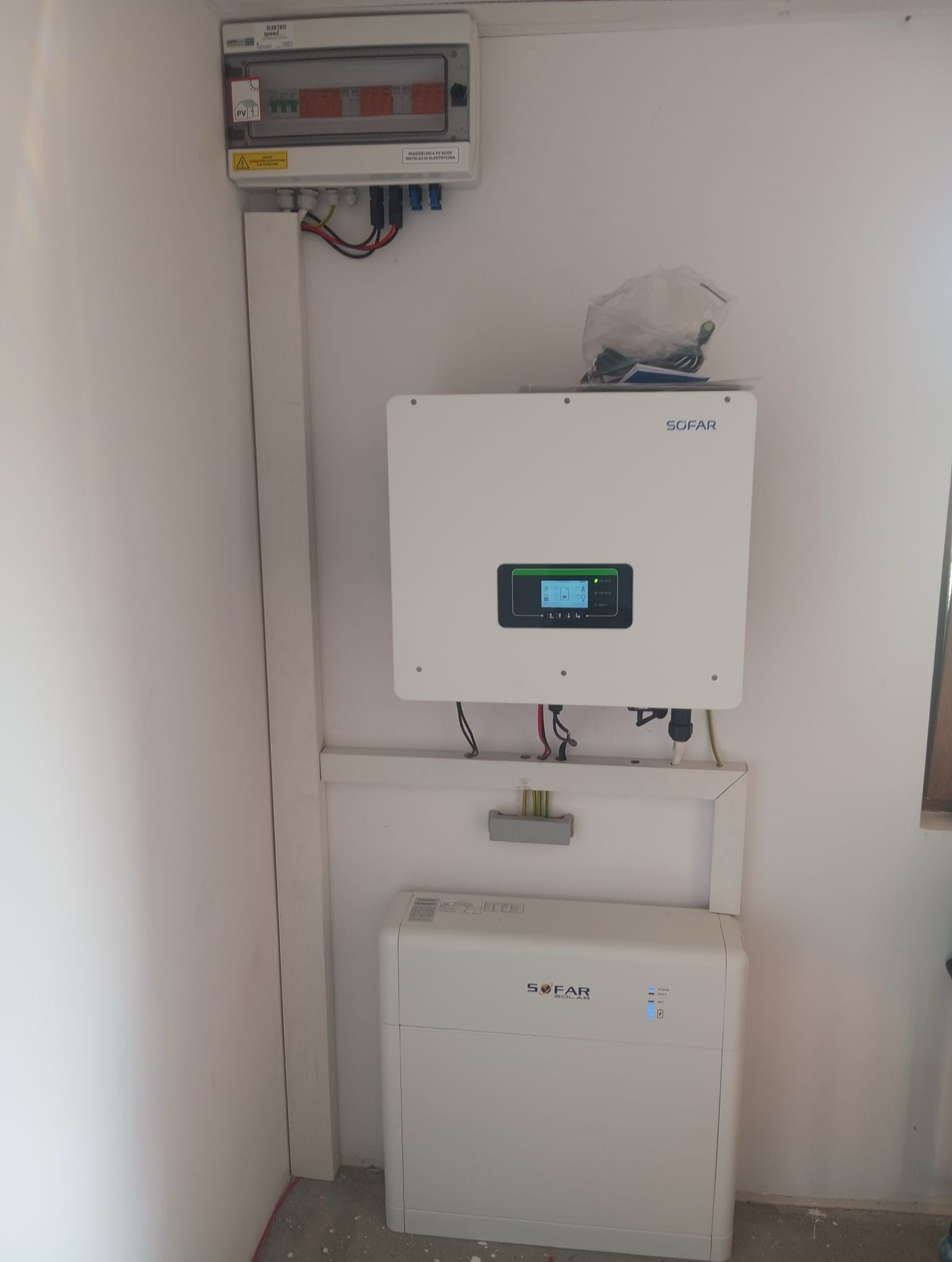 Biała skrzynka rozdzielcza PV, inwerter Sofar i akumulator na ścianie wewnątrz budynku. Widoczne kable i przewody. Instalacja fotowoltaiczna w trakcie montażu.