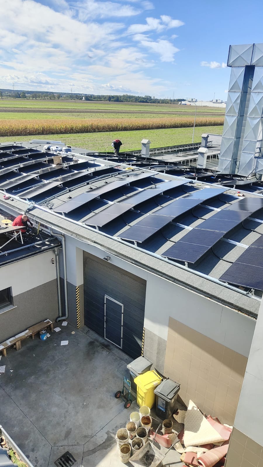 Montaż paneli fotowoltaicznych na dachu budynku przemysłowego, widok z góry na instalatorów pracujących na tle pól uprawnych i błękitnego nieba.