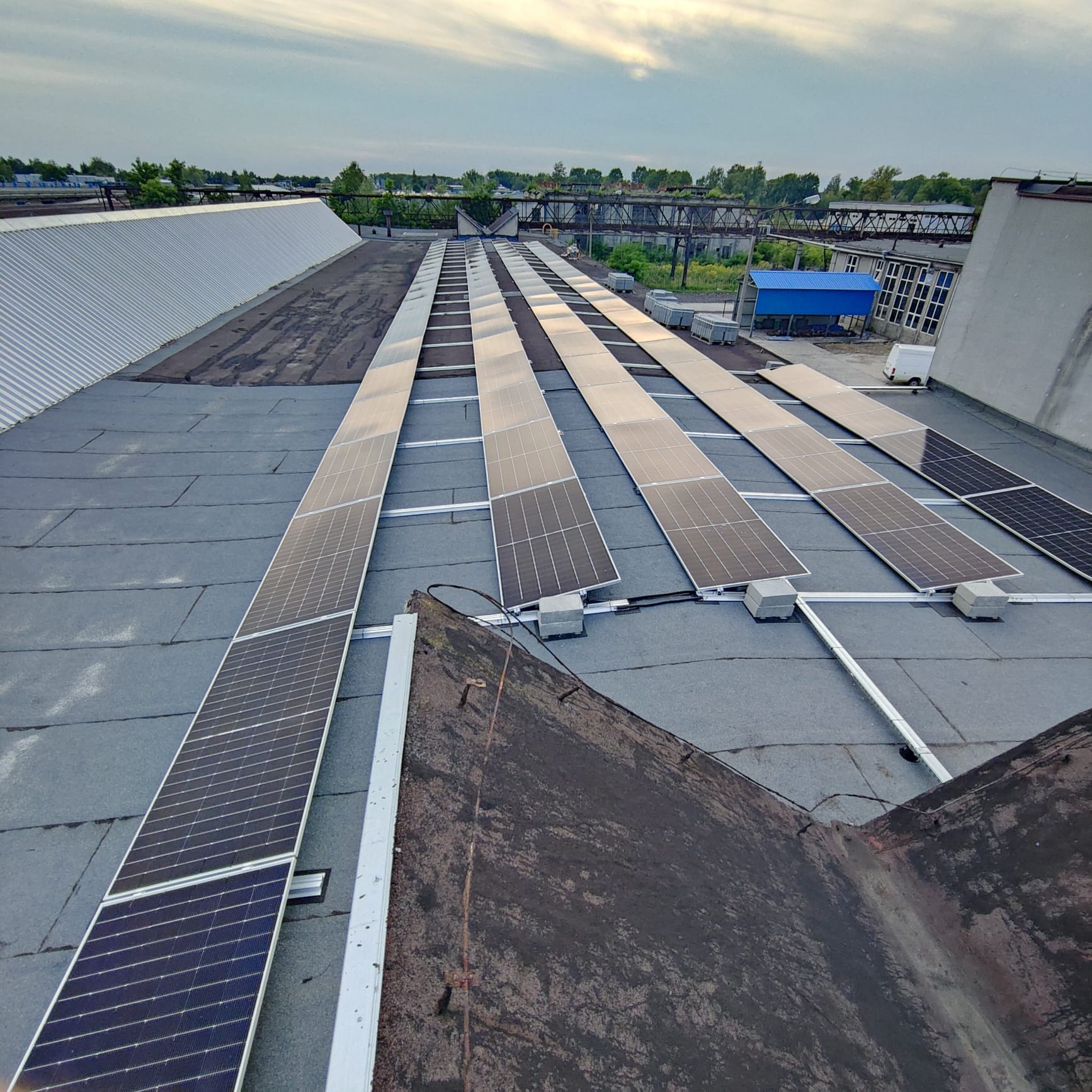 Panele fotowoltaiczne w trakcie montażu na płaskim dachu przemysłowym, widok z góry, częściowo zamontowane, w tle zabudowania przemysłowe i niebo.
