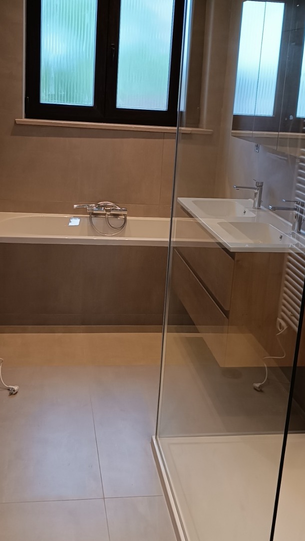 Nowoczesna łazienka z wanną, umywalką i kabiną prysznicową oddzieloną szklaną ścianką. Beżowe płytki na ścianach i podłodze. Minimalistyczny design.