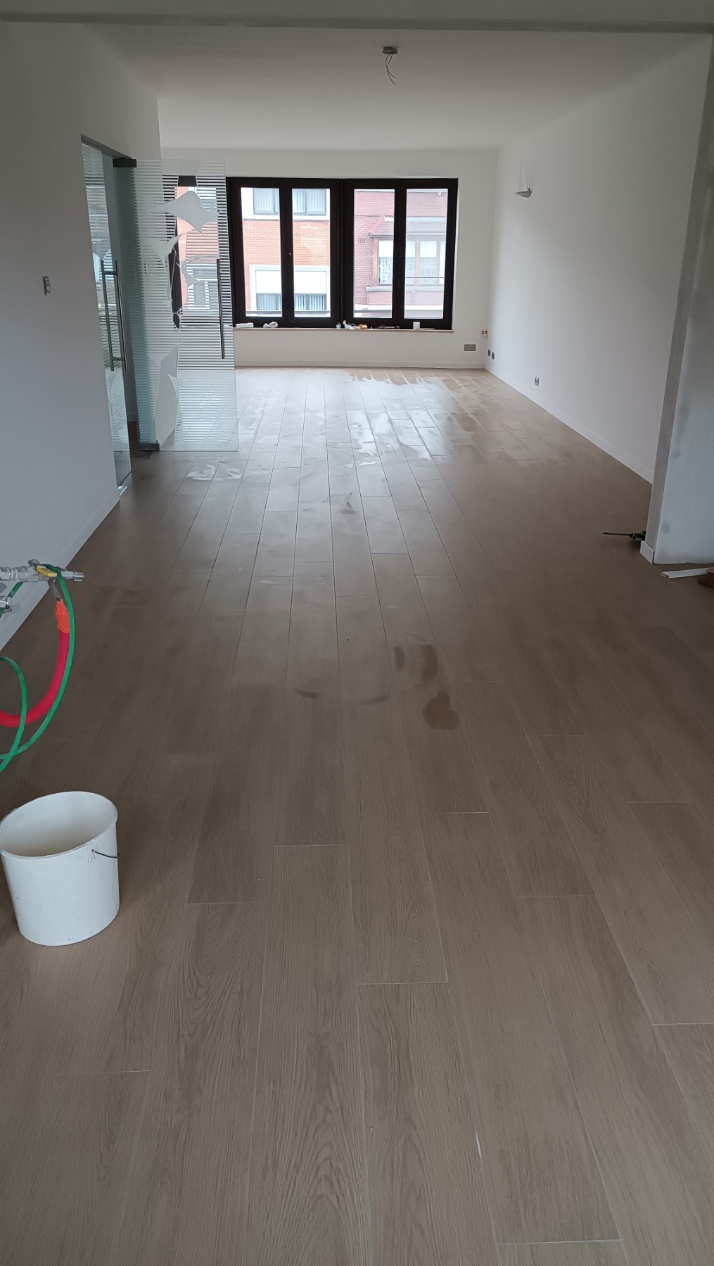 Jasne panele podłogowe w pustym pokoju z dużym oknem i grzejnikiem. Widoczne narzędzia i wiadro, sugerujące prace wykończeniowe. Minimalistyczny wystrój.