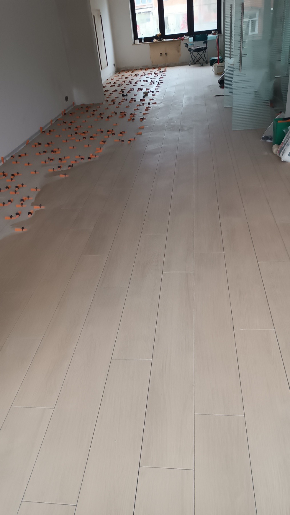 Ułożona podłoga z płytek w jasnym odcieniu, z pomarańczowymi krzyżykami dystansowymi wzdłuż ściany, widok z perspektywy podłogi, wnętrze w trakcie wykańczania.