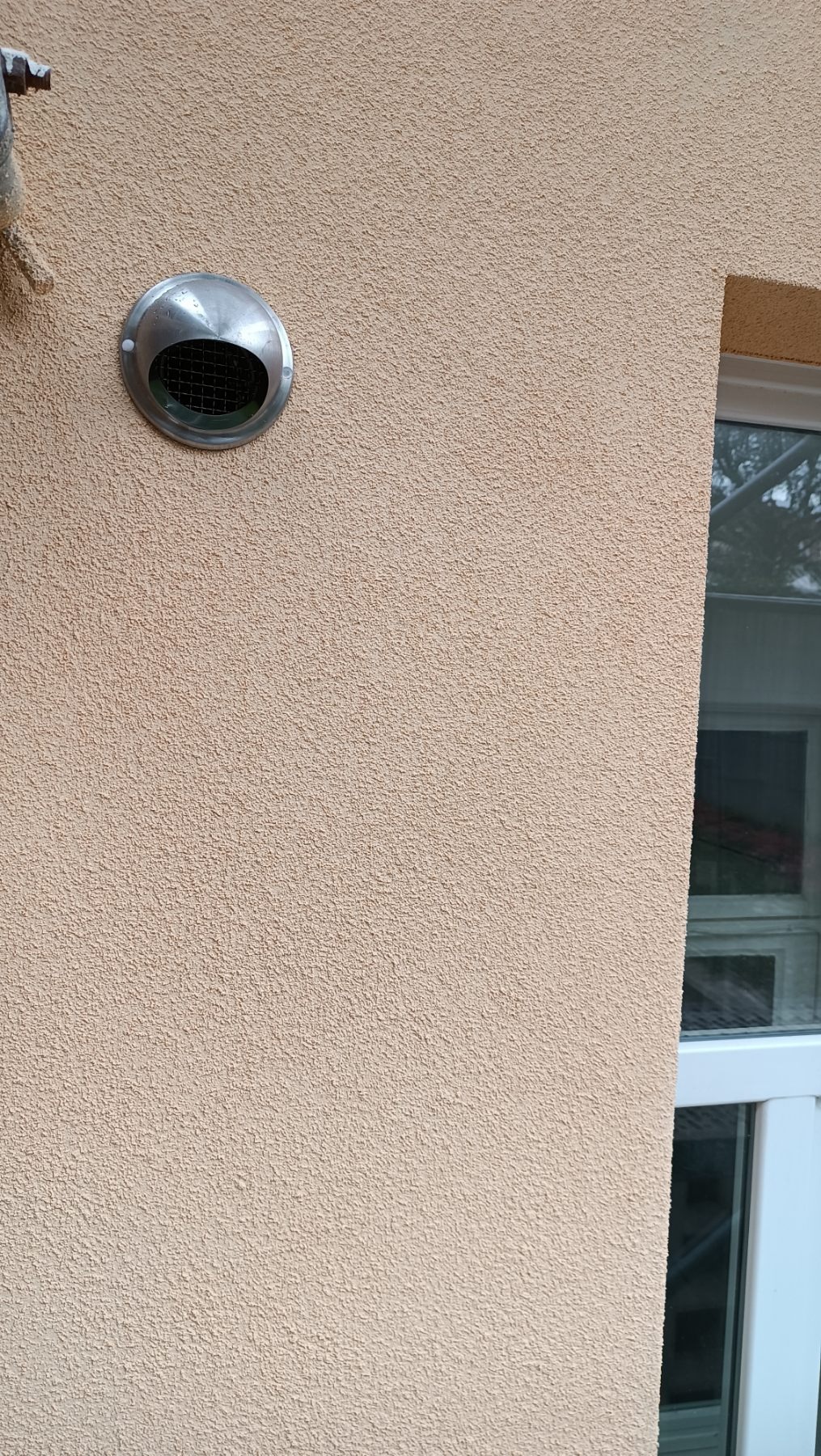 Beżowa elewacja z oknem i metalową kratką wentylacyjną. Chropowata faktura tynku, widoczne detale okna i siatka wentylacyjna. Ujęcie z bliska, naturalne światło.