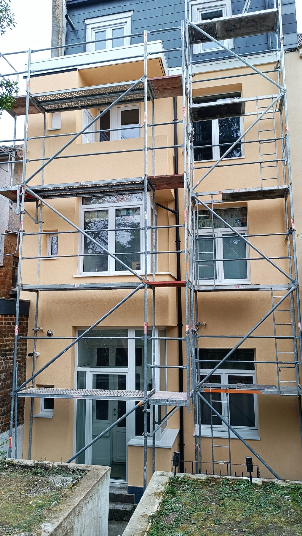 Elewacja budynku w kolorze ciepłego beżu, z białymi oknami i drzwiami, otoczona rusztowaniem. Widoczne detale wykończenia i odnowiona fasada.