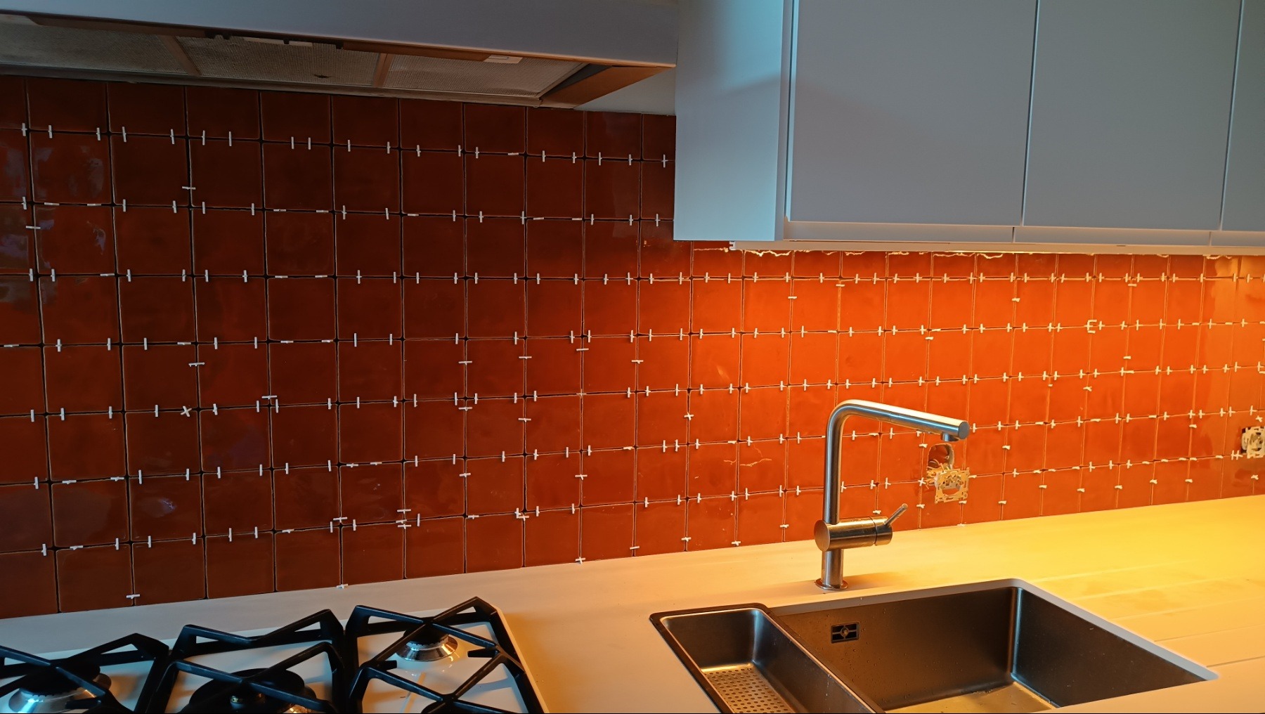 Ściana w kuchni z brązowymi, błyszczącymi płytkami w trakcie układania. Widoczne krzyżyki dystansowe, bateria kuchenna i fragment kuchenki gazowej.