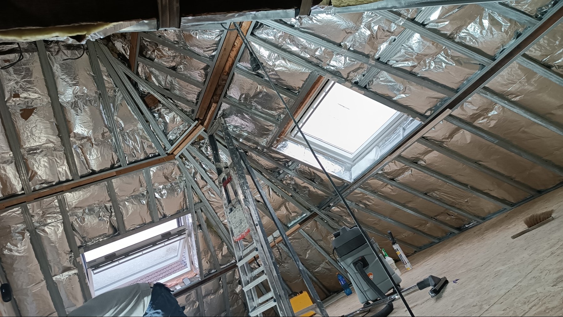 Srebrna izolacja poddasza z metalową konstrukcją i oknami dachowymi. Widoczny drabina i odkurzacz. Ujęcie z dołu, szeroki kąt.
