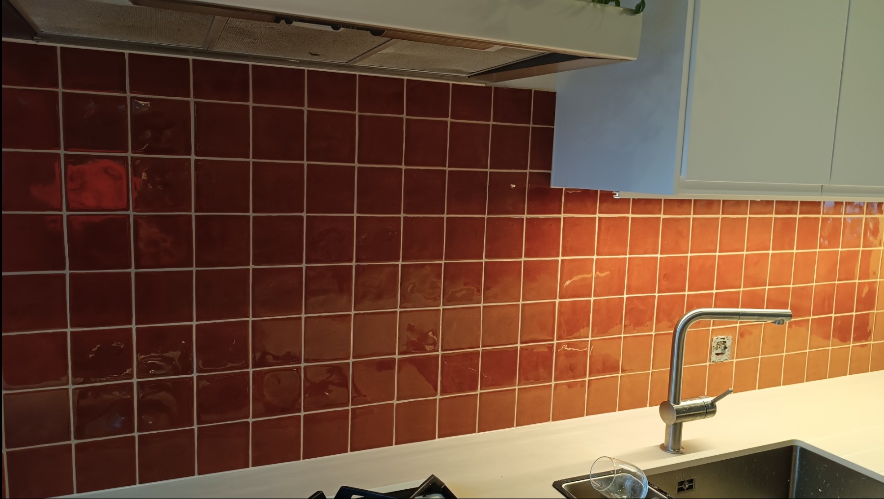 Błyszczące, terakotowe płytki kwadratowe na ścianie nad blatem kuchennym, z widocznym fragmentem zlewu i baterii. U góry białe szafki i okap.