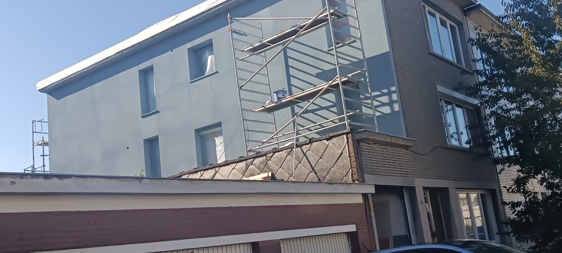 Świeżo pomalowana, błękitna elewacja budynku z szarym fragmentem, widoczne rusztowania i wiadro z farbą. Częściowo widoczny dach i drzewo.