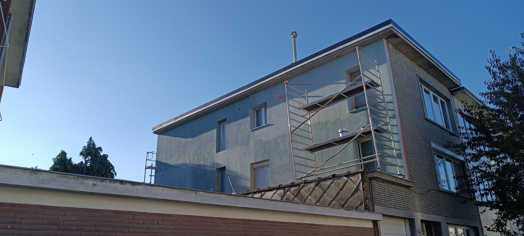 Dwupiętrowy budynek w trakcie malowania elewacji, z rusztowaniem przy ścianie. Widoczne okna zabezpieczone folią i puszka z farbą. Jasne, błękitne niebo w tle.