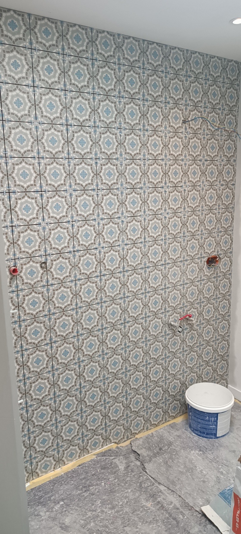 Ściana wyłożona dekoracyjnymi, wzorzystymi płytkami w odcieniach szarości i błękitu, z widocznymi krzyżykami dystansowymi i przygotowanymi punktami instalacji wodnej.