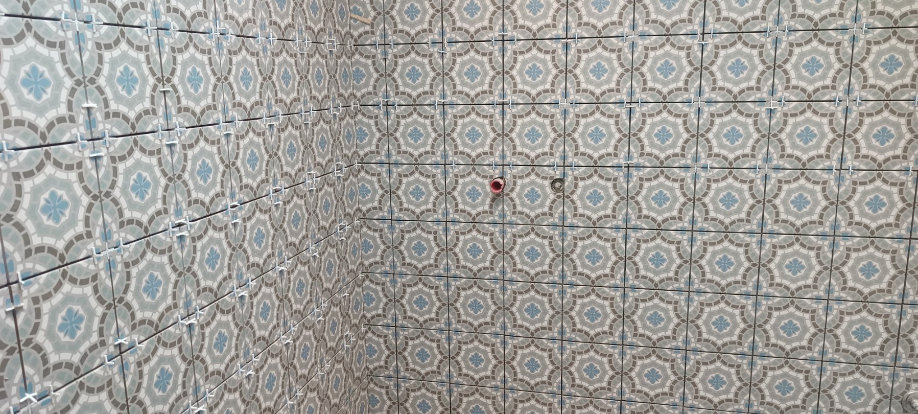 Ściana wyłożona dekoracyjnymi, kwadratowymi płytkami w odcieniach szarości i błękitu, z widocznymi krzyżykami dystansowymi i otworami na instalacje.