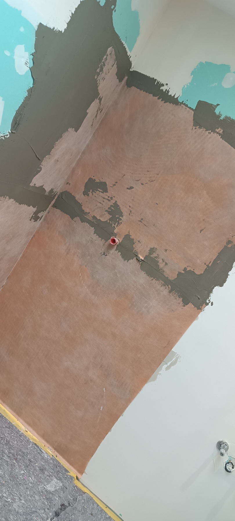 Ściana w trakcie prac wykończeniowych: częściowo zagruntowana na brązowo, z widocznymi fragmentami szarej masy szpachlowej i niebieskiej farby. Widoczne gniazdko elektryczne.