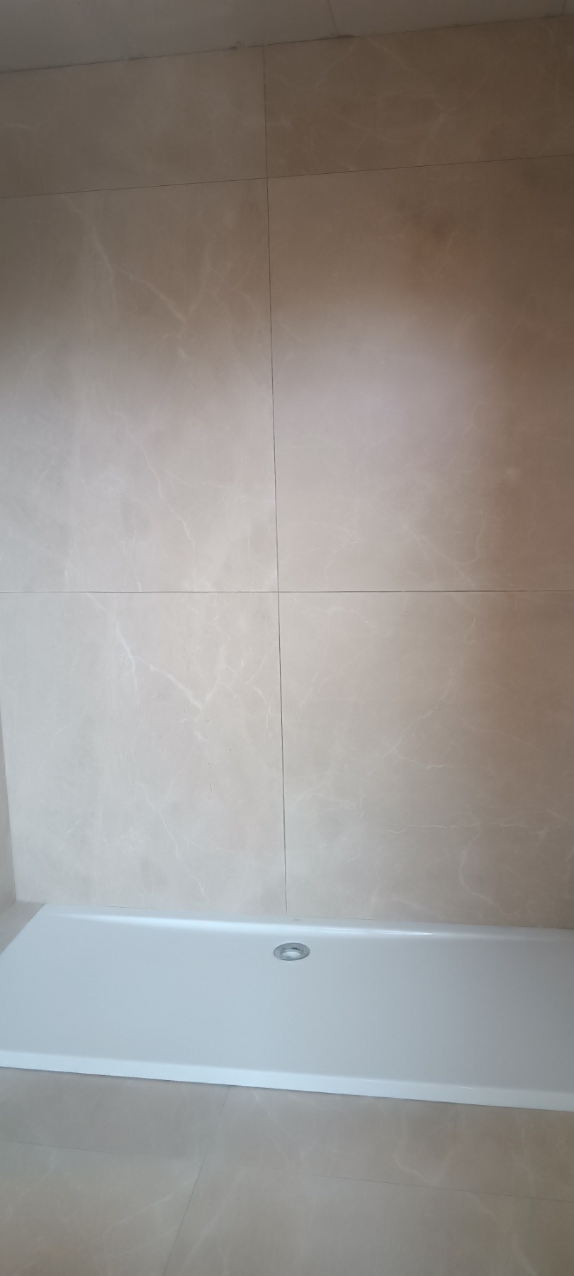 Beżowe płytki imitujące marmur na ścianie i podłodze łazienki, z widocznym białym brodzikiem z odpływem. Minimalistyczny design, ciepłe odcienie.