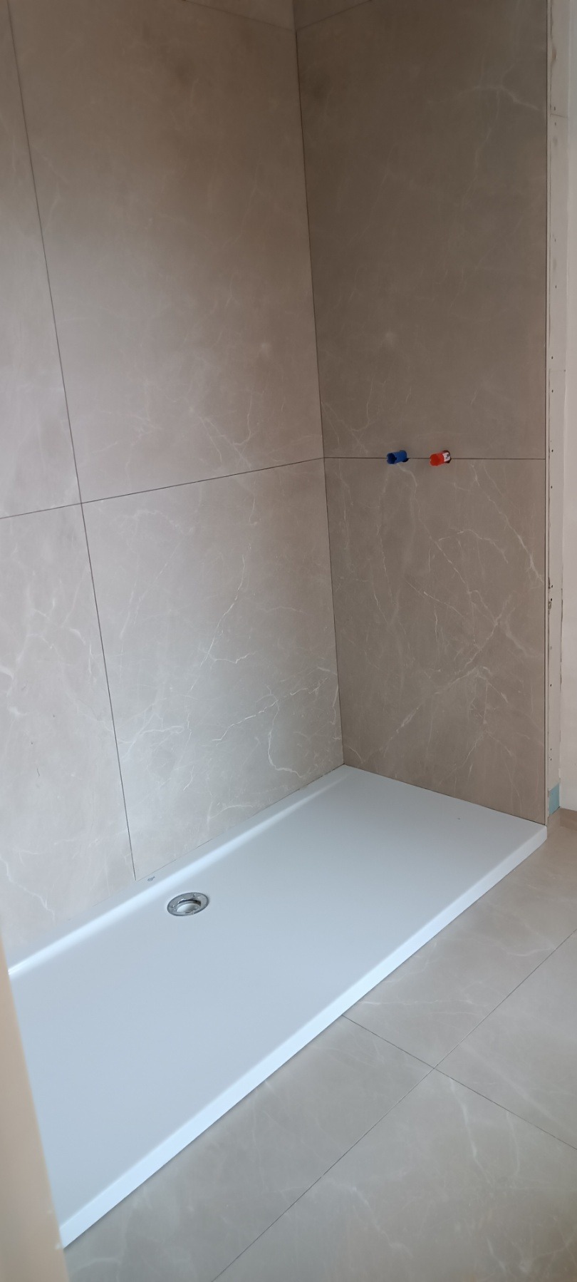 Wykończona kabina prysznicowa z beżowymi płytkami imitującymi marmur i białym brodzikiem. Widoczne przyłącza wodne.