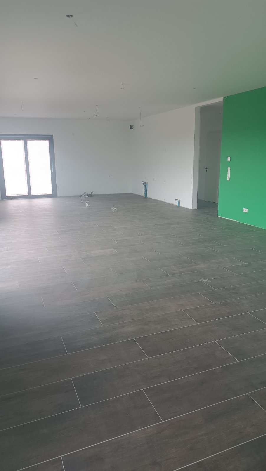 Ułożona, ciemna podłoga z płytek imitujących drewno w przestronnym, jasnym pomieszczeniu z zieloną ścianą. Widoczne wyprowadzenia instalacji.