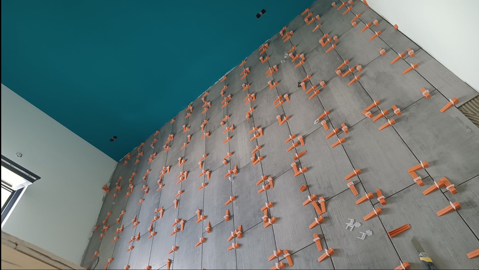 Ściana w trakcie układania szarych płytek z użyciem pomarańczowych klipsów poziomujących. Widoczny fragment turkusowego sufitu i biała ściana boczna. Ujęcie z dołu.