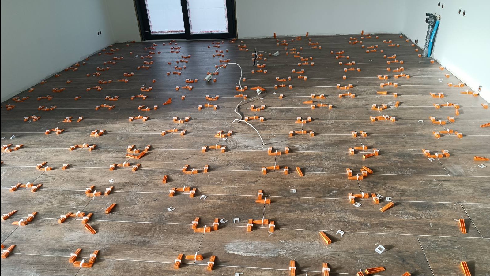 Układanie brązowych płytek podłogowych z użyciem pomarańczowych klipsów poziomujących i białych klinów w jasnym pomieszczeniu z oknem balkonowym.