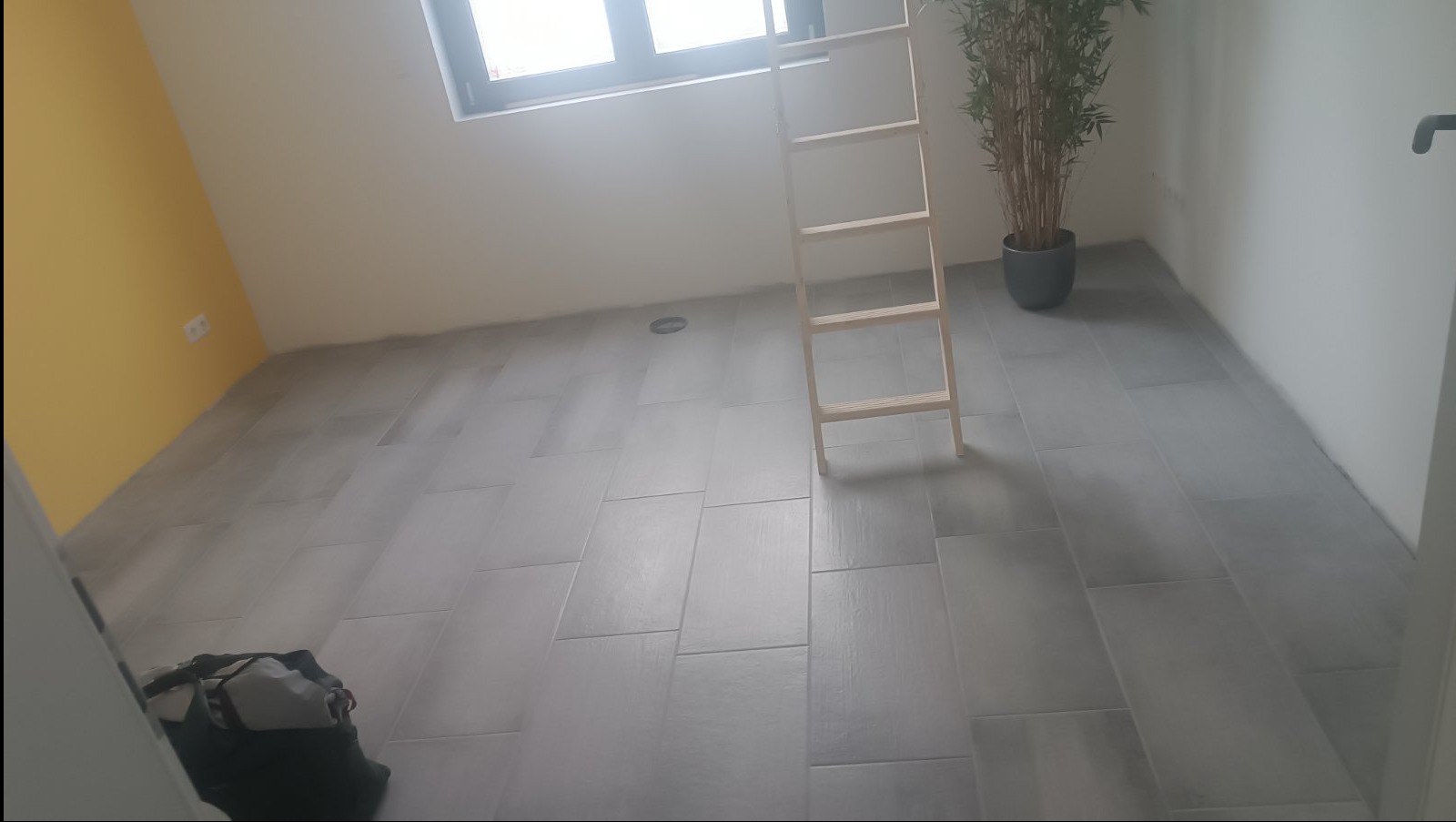 Ukończona podłoga z szarych, prostokątnych płytek w jasnym pokoju z żółtą ścianą. Widoczna drabina i roślina w doniczce. Torba narzędziowa w rogu.
