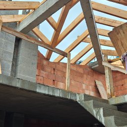 TP - BUD TOMASZ PIĄTEK - Surowy stan budynku w trakcie budowy: ściany z czerwonej cegły, betonowe schody i drewniana konstrukcja dachu pod błękitnym niebem. Widok z dołu.