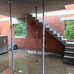 TP - BUD TOMASZ PIĄTEK - Wnętrze w trakcie budowy: murowane ściany z pustaków ceramicznych, betonowe schody, podpory stropowe, okna z widokiem na zieleń. Częściowo zalana posadzka.