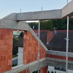 TP - BUD TOMASZ PIĄTEK - Wznoszenie ścian z czerwonej cegły na budowie domu jednorodzinnego, wsparte metalowymi stemplami. Widoczne betonowe nadproża i fragmenty konstrukcji. Tło: zabudowa mieszkalna.